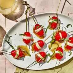 Szklane kieliszki z napojem i talerz z przekąskami na patyczkach: pomidorki koktajlowe z mozzarellą i bazylią.