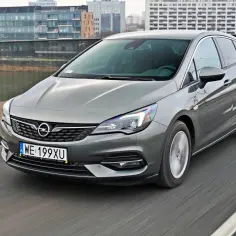 Szary Opel Astra Turbo pędzi po moście, w tle nowoczesne budynki.