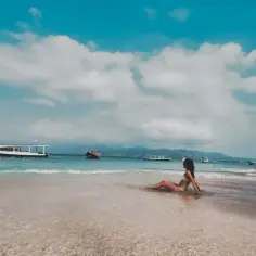 Kobieta w bikini siedzi w płytkiej wodzie na plaży, patrząc na morze z łodziami. To prawdziwy raj na ziemi.