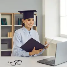 Uśmiechnięta absolwentka w togi, trzymająca teczkę, siedzi przy biurku z laptopem. Czy z licencjatem można pracować w szkole? Tak, to dobry początek kariery!