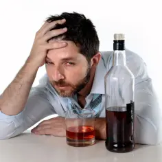 Mężczyzna z ręką na czole, z czerwonymi oczami, obok butelki i szklanki z alkoholem. Wygląda na to, że cierpi z powodu zatrucia alkoholowego.