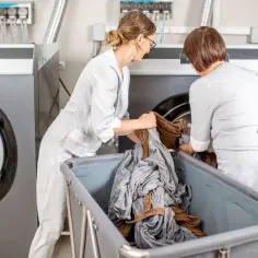 Pracownice pralni chemicznej w białych fartuchach wkładają pranie do profesjonalnych pralek. To pokazuje, dlaczego profesjonalne pralnie chemiczne nie używają zwykłych proszków do prania – potrzebują specjalistycznych środków.