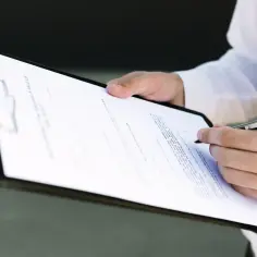 Dłoń podpisuje dokument, być może umowę kupna-sprzedaży nieruchomości. W tle widać fragment białej koszuli. Warto sprawdzić, czy nie ma tam ukrytych informacji o vat-r.