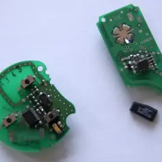 Dwa zielone układy elektroniczne, jeden okrągły, drugi podłużny, z widocznymi elementami i chipem. Obok leży mały czarny chip, prawdopodobnie kluczowy element immobilizera.