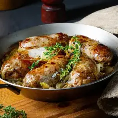 Pieczona w piekarniku biała kiełbasa surowa z cebulą, podana w metalowej patelni z gałązkami tymianku.
