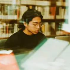 Student w okularach pochylony nad laptopem w bibliotece, przygotowuje podanie o indywidualny tok studiów uzasadnienie przykład.
