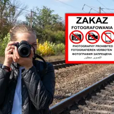 Kobieta z aparatem przy torach kolejowych, ignoruje znak "ZAKAZ FOTOGRAFOWANIA".