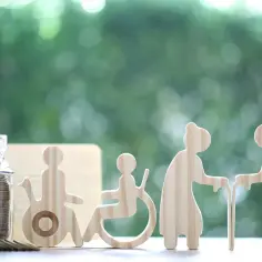 Skarbonka z monetami i drewniane figurki symbolizujące osoby w różnym wieku i niepełnosprawne. Czy świadczenie wspierające wlicza się do dochodu?