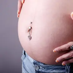 Brzuszek w ciąży z kolczykiem w pępku, ozdobionym serduszkiem. Dłoń z pierścionkami delikatnie dotyka skóry.