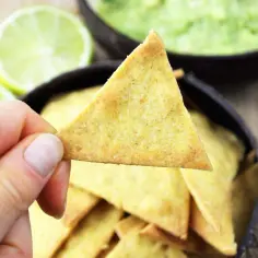 Dłoń trzyma nachos nad miską z guacamole, obok połówki limonki.