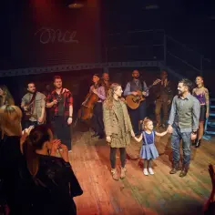 Aktorzy i muzycy z spektaklu "Once" na scenie, z małą dziewczynką trzymającą za rękę.