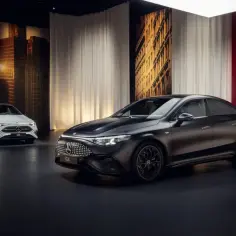 Dwa modele Mercedes CLA 2025: jeden biały, drugi grafitowy, prezentowane na tle miejskiej panoramy i czerwonej ściany z logo marki.