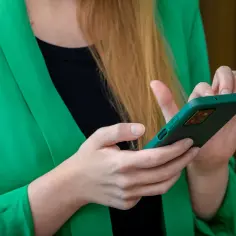 Kobieta w zielonej marynarce trzyma telefon. Szuka informacji, gdzie jest schowek w telefonie, by ukryć ważne dane.