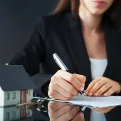 Rejent podpisuje dokumenty dotyczące zakupu domu. Obok leży model domu i klucze.