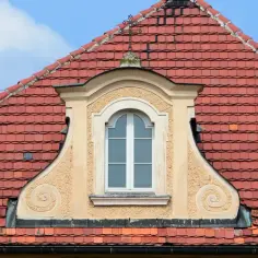 Eleganckie, małe okienko w dachu z ozdobnymi wolutami, otoczone czerwonymi dachówkami.