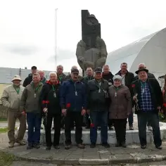 Grupa ludzi z czerwonymi goździkami przed pomnikiem i sarkofagiem elektrowni Czarnobyl.