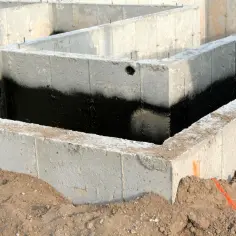 Surowe ściany fundamentowe z betonu, częściowo pokryte czarną izolacją przeciwwilgociową, czekają na dalsze prace budowlane.