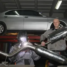 Mechanik spawa układ wydechowy, zastanawiając się, czy downpipe jest legalny. Srebrny samochód uniesiony na podnośniku.