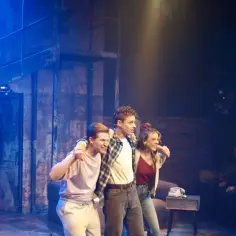 Trzech młodych ludzi na scenie, w tle ceglana ściana i niebieskie światła. Może to scena z musicalu "Tik Tik Bum" w Rzymie?