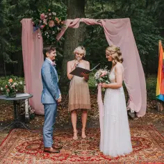 Ceremonia ślubna w lesie, gdzie można wziąć ślub homoseksualny. Dwie kobiety, jedna w garniturze, druga w sukni ślubnej, stoją przed urzędniczką. Tęczowa flaga symbolizuje miłość.
