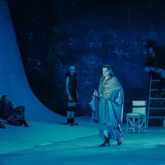 Scena teatralna w niebieskim świetle. Grupa aktorów w kostiumach, jedna postać w centrum w luźnej, połyskującej szacie. Czy to scena z jednego z wielu teatrów w Warszawie?