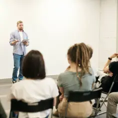 Mężczyzna z tabletem przemawia do grupy słuchaczy. Czy z licencjatem można uczyć w szkole? Pytanie wisi w powietrzu.