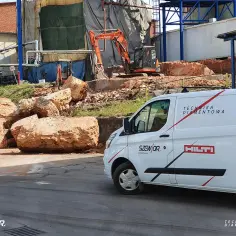 Wyburzenie ceglanych bloków i pytanie: ile kosztuje kucie betonu? Na placu budowy koparka i biały van z napisem "Technika Diamentowa".