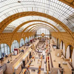 Wnętrze muzeum d'Orsay z imponującym szklanym sufitem i łukami. Zwiedzający spacerują wśród rzeźb i dzieł sztuki.