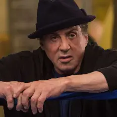 Sylvester Stallone jako Rocky Balboa, w czarnym kapeluszu i bluzie, opiera się o ring bokserski.