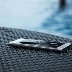 Smartfon z kroplami wody na ekranie, leżący na plecionej powierzchni obok basenu. To dowód, że IP68 co oznacza: ochronę przed zanurzeniem.