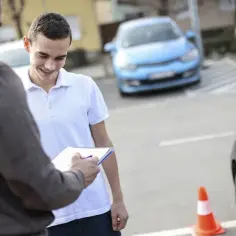 Młody mężczyzna z uśmiechem i aparatem na zębach słucha instruktora, który notuje coś na kartce. Czy to jego pierwsze kroki do zdobycia prawa jazdy?