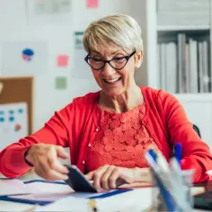 Uśmiechnięta starsza pani przy biurku sprawdza coś na telefonie, zastanawiając się, ile można dorobić do emerytury.