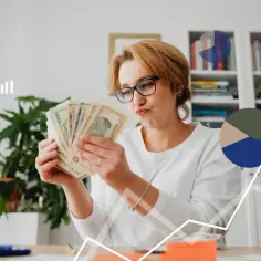 Kobieta z pieniędzmi zastanawia się, jak legalnie obniżyć wynagrodzenie pracownikowi w kryzysie. Wykresy sugerują trudną sytuację finansową.