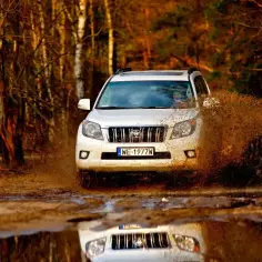 Biały Land Cruiser 100 pruje przez błotnistą kałużę w lesie, wzbijając fontannę brudu.