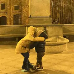 Dwoje dzieci w zimowych ubraniach przytulających się na tle ceglanej kamienicy i fontanny.