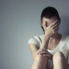 Kobieta siedzi przy ścianie, zakrywając twarz dłońmi. Może to symbolizować trudności związane z przyjmowaniem leków, jak np. rexetin skutki uboczne.
