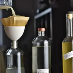Klarowne drinki metodą milk punch w domu: płyn przelewany przez filtr papierowy do butelki, obok stoją butelki z gotowymi miksturami.