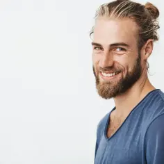 Uśmiechnięty mężczyzna z blond włosami spiętymi w kok i gęstą brodą bez wąsa.