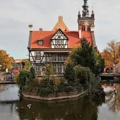 Jesień na gdańskiej starówce. Urokliwy dom o konstrukcji szachulcowej nad kanałem, w tle wieża ratusza i złote liście drzew.