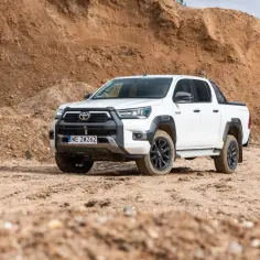Biały Toyota Hilux 2025 gotowy na terenowe wyzwania, zaparkowany na piaszczystym podłożu, z piaskowymi wzniesieniami w tle.