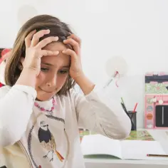Dziewczynka z rękami na głowie, wyrażająca stres u dziecka w wieku szkolnym. Na biurku leżą książki i przybory.