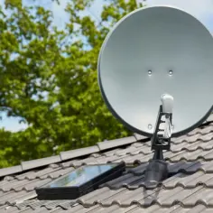 Srebrna antena satelitarna na szarym dachu, obok okna dachowego. W tle zielone drzewa i błękitne niebo.
