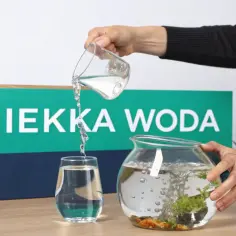 Osoba przelewa wodę do akwarium, pokazując, jak zmiękczyć wodę w akwarium. Na tle napis "MIĘKKA WODA".