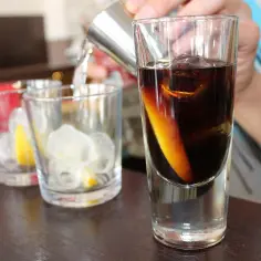 Przygotowanie drinka z whisky: ciemny płyn z plasterkiem pomarańczy w szklance, obok szklanki z lodem i cytryną.
