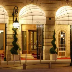 Eleganckie wejście do słynnego paryskiego hotelu Ritz, oświetlone nocą, z ozdobnymi markizami i spiralnymi drzewkami.