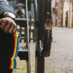 Dłoń trzyma tęczę, symbol dumy. Osoba w czarnych spodniach i szarej bluzie, z zegarkiem na nadgarstku, siedzi na schodach.