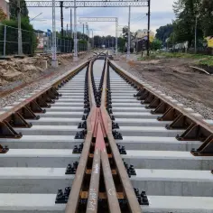 Nowe tory kolejowe, rozjazd i betonowe podkłady. W tle widać budynki i słupy trakcyjne. Akcje Torpol w budowie.