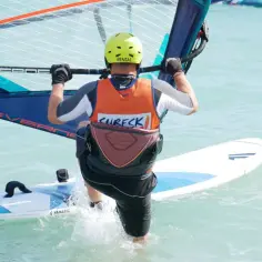 Osoba w kasku i kamizelce uprawia windsurfing na turkusowej wodzie.