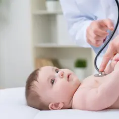 Lekarz osłuchuje dziecko, sprawdzając, czy nie ma szmerów nad sercem. Maluch spokojnie leży na białym podłożu.