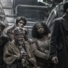 Grupa pasażerów Snowpiercer: Arka Przyszłości, w tym mężczyzna w czapce i płaszczu, siedzi w ciasnym wagonie.